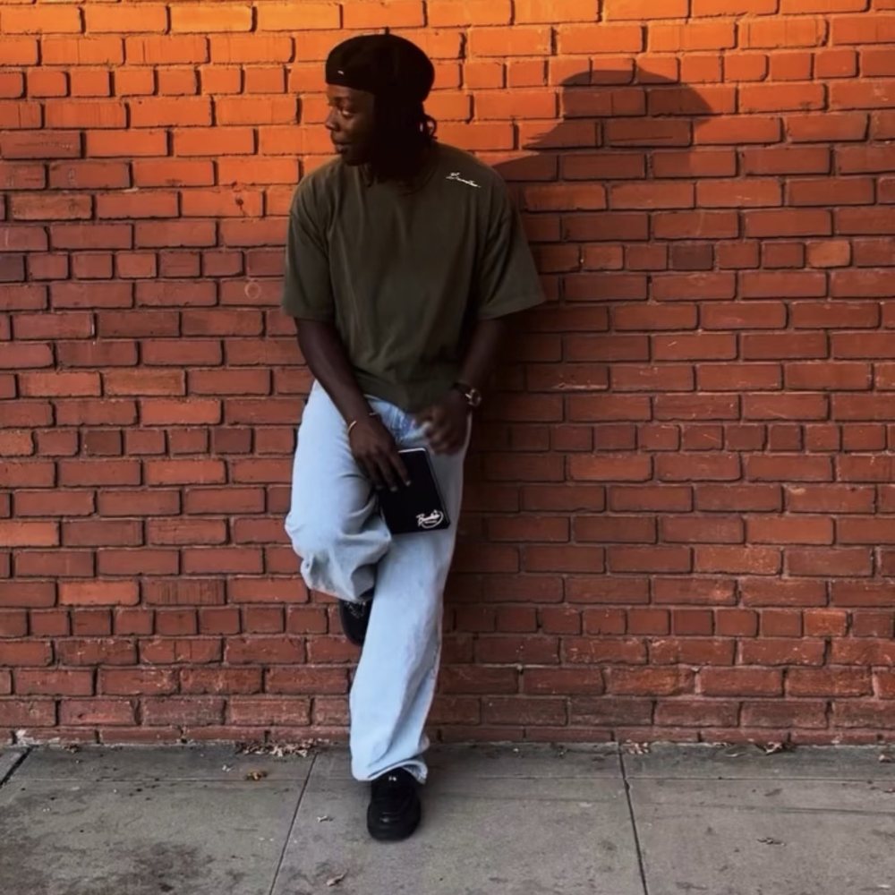 Raymell leaning on a red brick wall in an olive T-shirt with Boundless in script on the shoulder, holding a black notebook