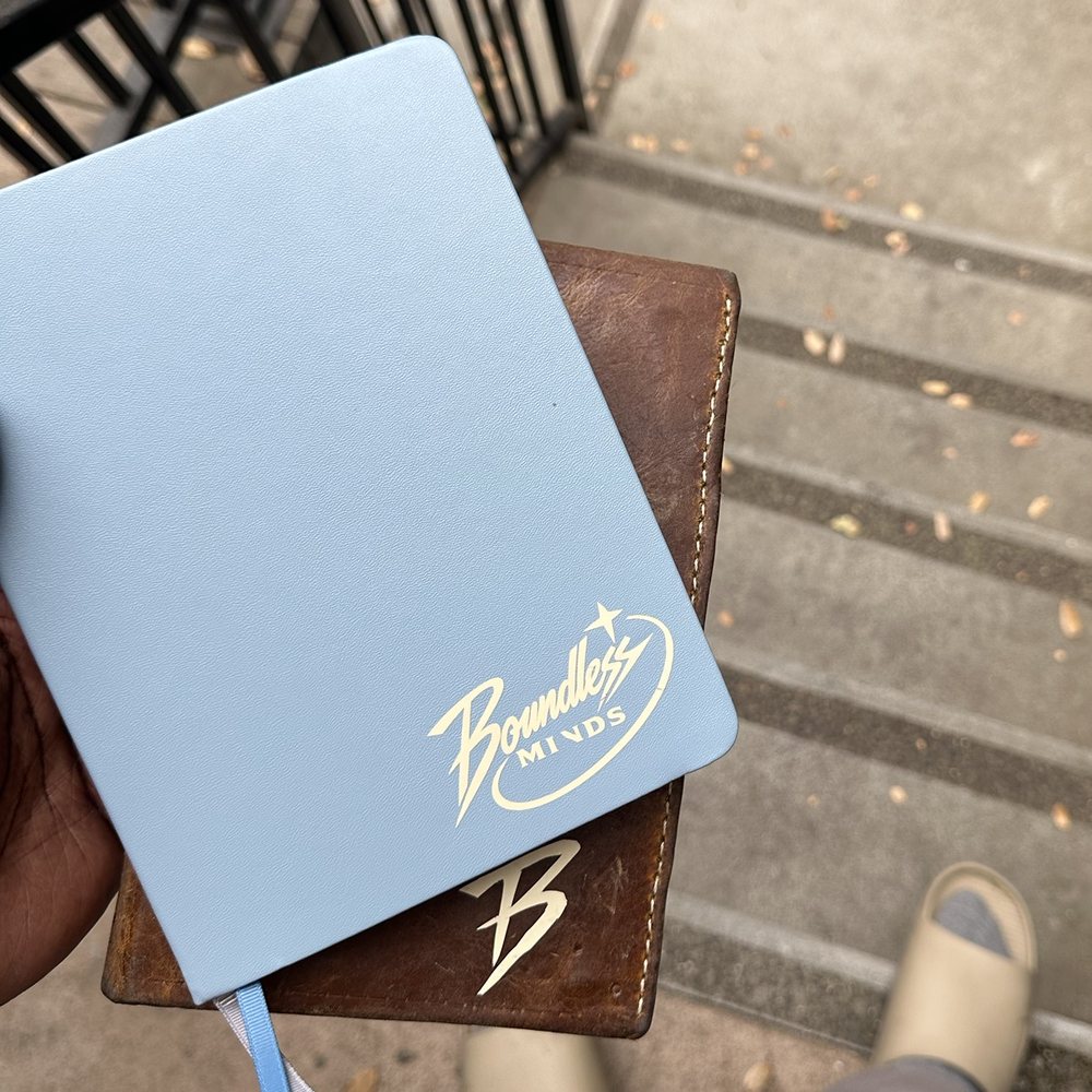 A light blue notebook with the Boundless Minds logo in cream, held over a brown leather journal with a B on it