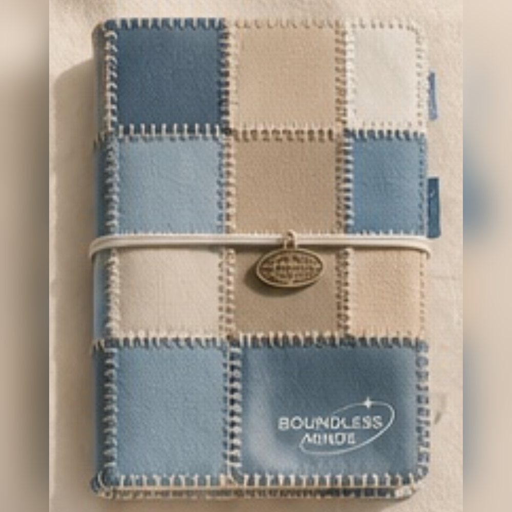A notebook with a patchwork cover of blue and tan squares, stitched edges, an elastic strap with a round charm, and a Boundless Minds logo